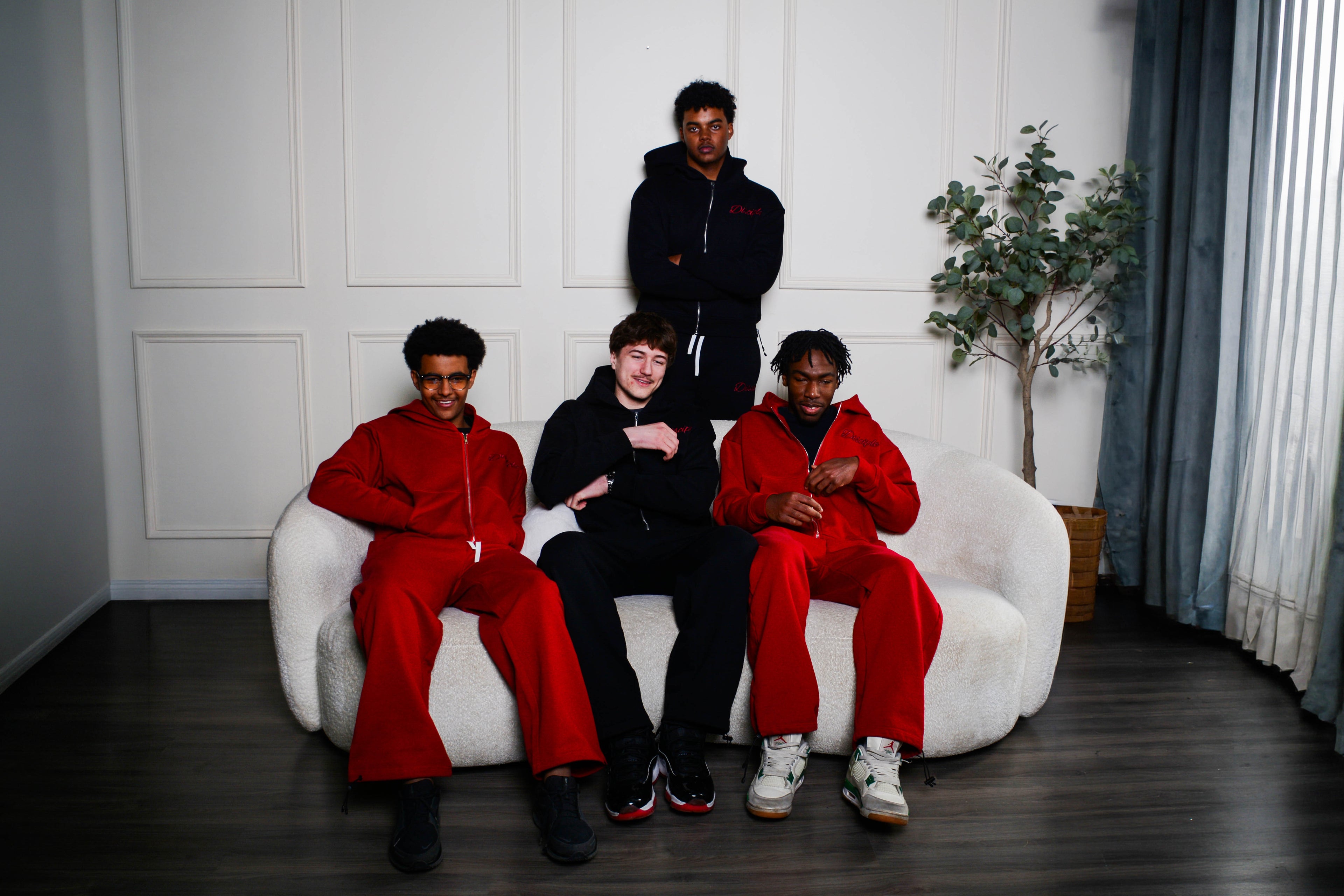 Four individuals in red and black outfits sitting on a white couch in a room with a plant and curtains.